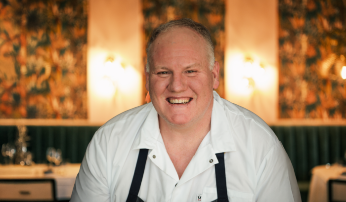 Chef-partner James Trees smiles inside Bar Boheme, wearing a white chef's coat and dark apron, with the restaurant's warm, tropical-inspired dining room softly blurred in the background.