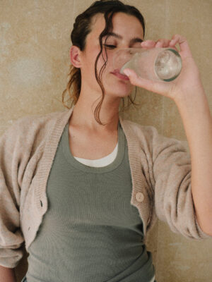 A person wearing a sage green ribbed tank top and a light beige cardigan drinks a glass of water indoors. Their dark hair is loosely pulled back with soft curls framing their face, while the warm neutral background and relaxed pose create a calm, wellness-focused atmosphere.