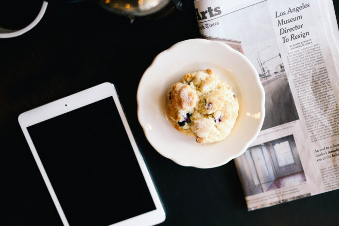 A fresh blueberry scone sits on a white plate beside a folded newspaper and a tablet on a dark café table, suggesting a relaxed morning of reading over breakfast.