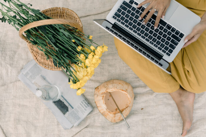 An overhead view of a person working on a laptop while seated on a light-colored blanket. A woven basket filled with yellow flowers, a newspaper, a glass bottle of water, and a fresh coconut with a metal straw create a bright, relaxed remote-work setting.