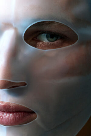 Close-up portrait of a person wearing a translucent white sheet face mask. One blue-green eye is visible through the mask's opening, while soft light highlights the skin and glossy lips. The tightly cropped image emphasizes the skincare treatment, creating a calm, intimate spa-like atmosphere.