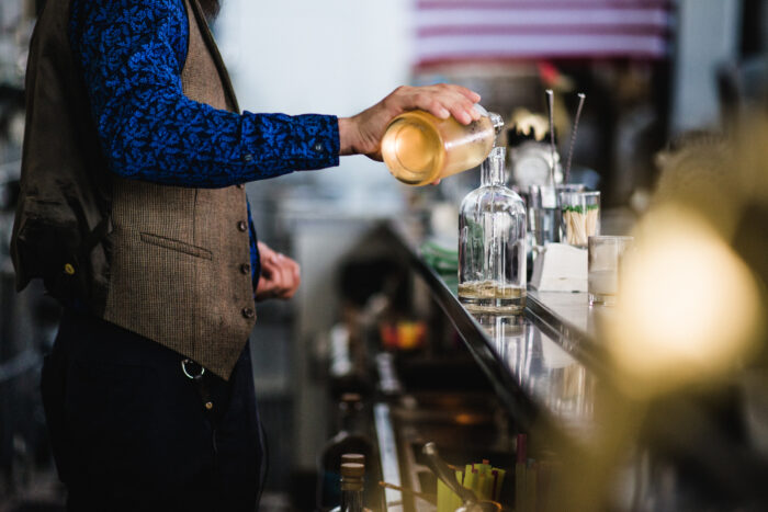 A bartender pours a freshly shaken cocktail from a metal shaker into a glass bottle behind a bar. Glassware, bar tools, and cocktail ingredients line the counter, while the softly blurred background creates the atmosphere of a busy craft cocktail bar.