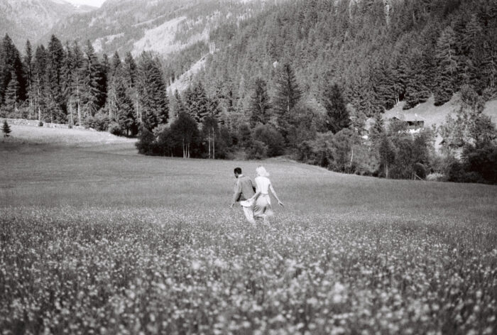 A black-and-white photograph of a couple walking hand in hand through a wildflower meadow, surrounded by towering pine forests and mountains. Seen from behind, they move toward the wooded hillside, creating a quiet, timeless scene of romance and escape.