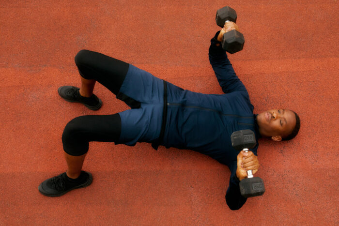 Overhead view of a person performing a strength-training exercise on an outdoor rubber track while holding two dumbbells. Wearing a navy athletic top, blue shorts, black leggings, and black training shoes, they are in a bridge position with one arm extended upward, highlighting balance, core strength, and functional fitness.