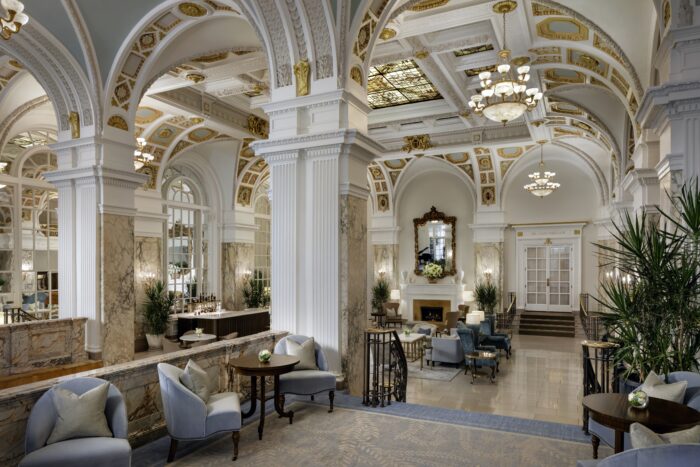 The grand lobby of The Hermitage Hotel in Nashville, featuring soaring marble columns, ornate gold-accented vaulted ceilings, crystal chandeliers, elegant seating areas, and a stately fireplace that reflects the hotel's Beaux-Arts architecture and historic grandeur.