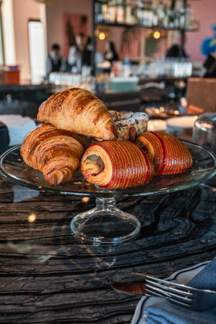 A glass pedestal displays an assortment of freshly baked pastries, including buttery croissants and fruit-filled laminated pastries, on the marble bar at Bar Boheme.