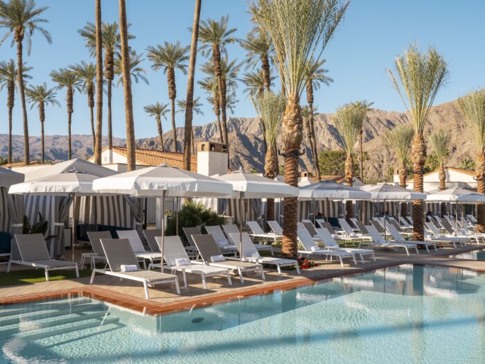 Sunlit pool at La Quinta Resort & Club featuring white lounge chairs, striped cabanas, tall palm trees, Spanish-style casitas, and the rugged Santa Rosa Mountains rising in the background under a clear blue sky.