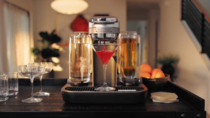 A Bartesian automated cocktail machine dispenses a bright red cocktail into a martini glass on a kitchen countertop. Bottles of spirits, glassware, and citrus fruit surround the machine in a warm, contemporary home setting.
