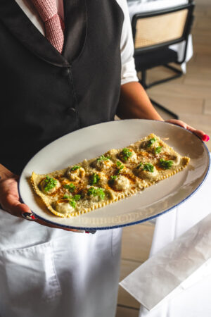 A server presents a large sheet of Raviole du Dauphiné—delicate French ravioli topped with browned butter, grated Parmesan, cracked black pepper, and fresh herbs—at Bar Boheme.