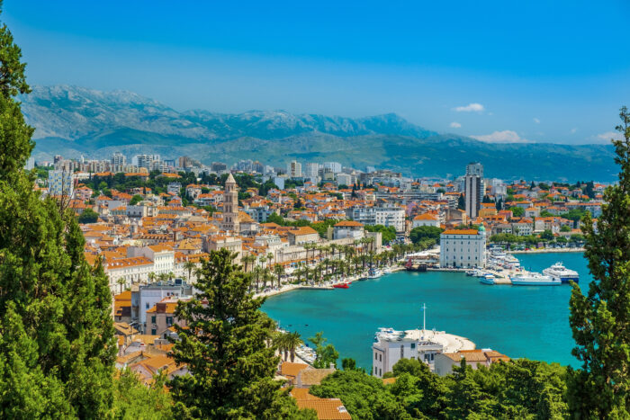 Panoramic view of Split in Dalmatia, Croatia with a view of terracotta rooftops, a palm-lined waterfront, and a marina filled with boats along a turquoise harbor. Framed by lush green trees in the foreground, the city stretches toward rugged mountains beneath a clear blue sky.
