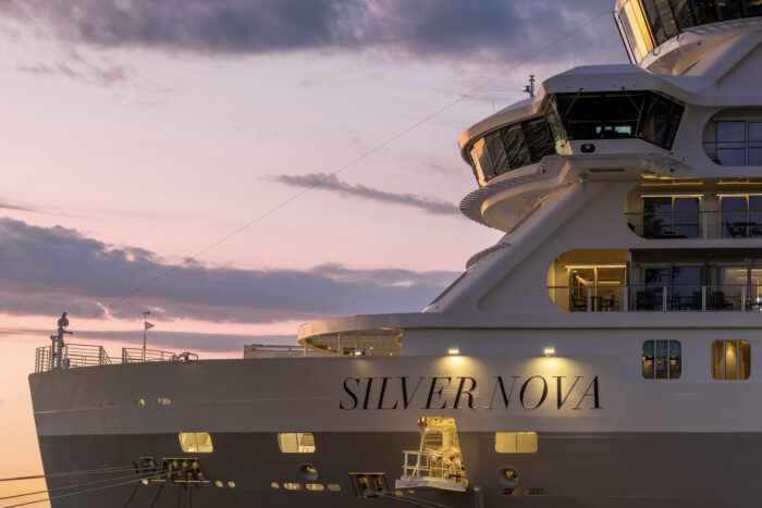 The bow and upper decks of the luxury cruise ship Silver Nova are illuminated at sunset, with the vessel's name prominently displayed on the hull. Warm interior lights glow through expansive windows as soft pink and purple clouds fill the evening sky above the docked ship.