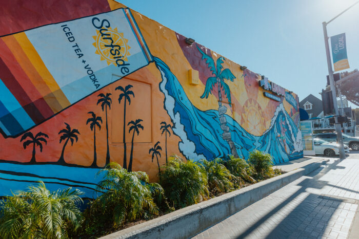Colorful tropical mural covers the exterior wall of a liquor store, featuring a giant Surfside iced tea and vodka can pouring into a curling ocean wave, with palm trees, a surfer, and a bright sunset. The vibrant street-corner scene is framed by landscaped greenery under a clear blue sky.