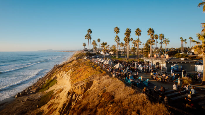 Golden-hour view of a bustling oceanfront restaurant perched atop coastal bluffs overlooking the Pacific Ocean. Palm trees line the shoreline while diners gather on the outdoor patio, with waves rolling onto the beach below beneath a clear blue sky.