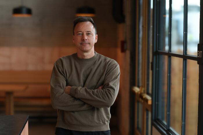 Portrait of chef Eric Bost standing with his arms crossed inside a warmly lit restaurant. Wearing an olive crewneck sweatshirt, he faces the camera with a calm, confident expression beside floor-to-ceiling windows, while softly blurred pendant lights and wooden seating create an inviting backdrop.