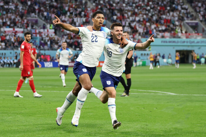 Two England soccer players celebrate a goal during a FIFA World Cup match, running across the field with their arms outstretched and pointing toward the crowd. A packed stadium of fans fills the background, while opposing players in red walk away as the celebration unfolds.