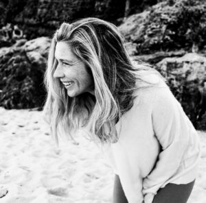 Black-and-white portrait of a smiling person with long, windswept hair leaning forward on a sandy beach. Wearing a light-colored knit sweater and looking off to the side, they appear to be laughing naturally. Rocky cliffs in the softly blurred background add texture and depth to the candid outdoor scene.