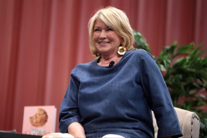 Martha Stewart smiles while seated on stage during a live interview, wearing a blue blouse and large gold earrings with a lapel microphone clipped to her neckline.