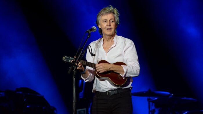 Paul McCartney performs onstage under blue lights, singing into a microphone while playing a ukulele during a live concert.