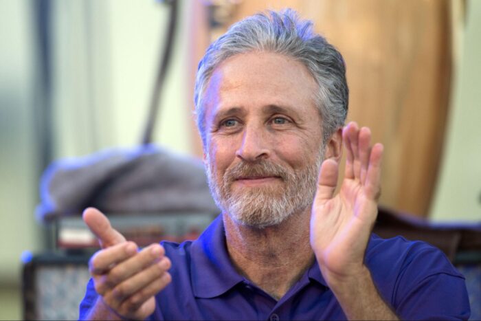 Jon Stewart smiles and applauds while seated at an event, wearing a purple polo shirt against a softly blurred indoor background.