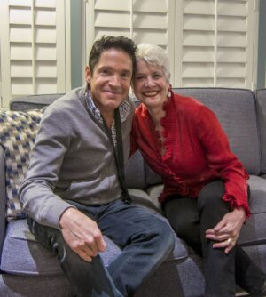 Dave Koz and Marsala Rypka sit closely together on a gray sofa, smiling warmly at the camera in a cozy indoor setting. The person on the left wears a gray zip-up sweater over a checkered shirt with dark pants, while the person on the right wears a bright red ruffled blouse with black pants and silver earrings. White plantation shutters fill the background, and a patterned accent pillow rests on the sofa beside them.