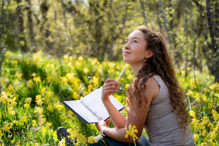 Thoughtful young woman holds a notebook and pen while practicing mindfulness in nature. Girl sits in the blooming forest, pondering her thoughts.