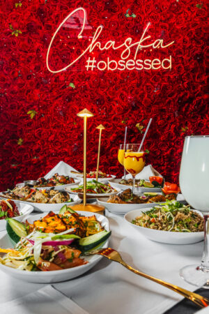 A table filled with Indian dishes and drinks set against Chaska’s vivid red rose wall with a glowing “Chaska #obsessed” sign.