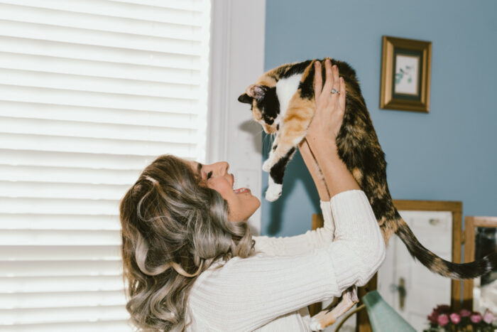 A woman smiles as she lifts a calico cat into the air inside a cozy home. Bathed in soft natural light, the affectionate moment captures the close bond between pet and owner, highlighting companionship, joy, and everyday connection.