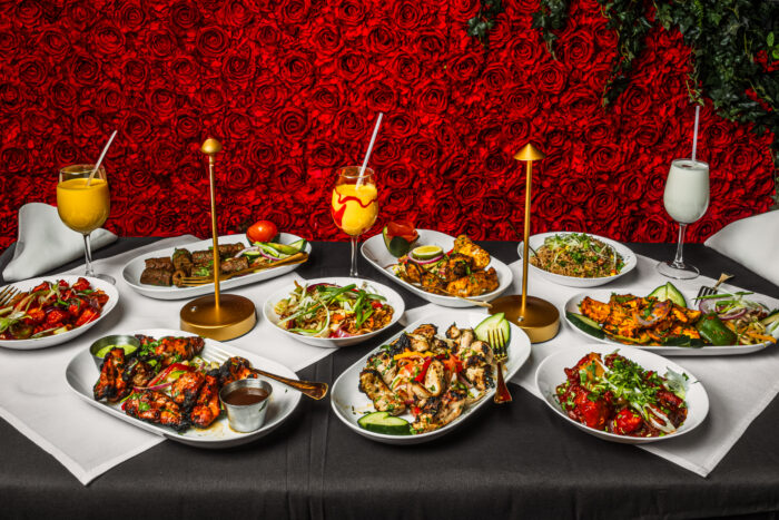 A table laden with colorful Indian dishes, grilled meats, rice and drinks, framed by gold table lamps and a dramatic wall of red roses.