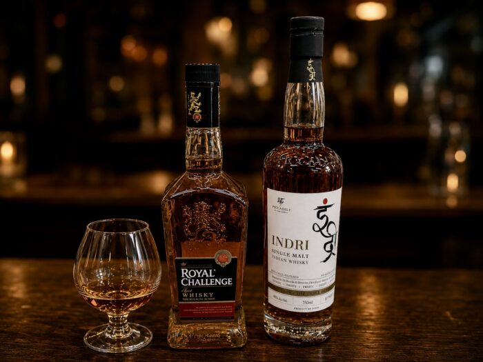 Bottles of Indri single malt Indian whisky and Royal Challenge whisky displayed beside a glass of amber whisky on a warmly lit bar.