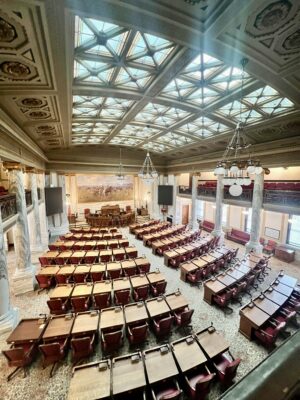 Interior of the Montana State Capitol legislative chamber featuring marble columns, skylights and rows of lawmakers’ desks.