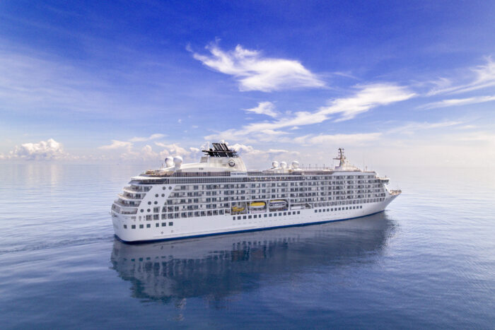 The World residential mega-yacht sailing on calm blue waters beneath a bright, cloud-filled sky.