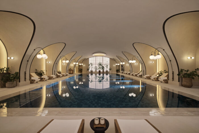 Indoor swimming pool at Six Senses London, framed by curved architectural details, lounge chairs, greenery and soft lighting.