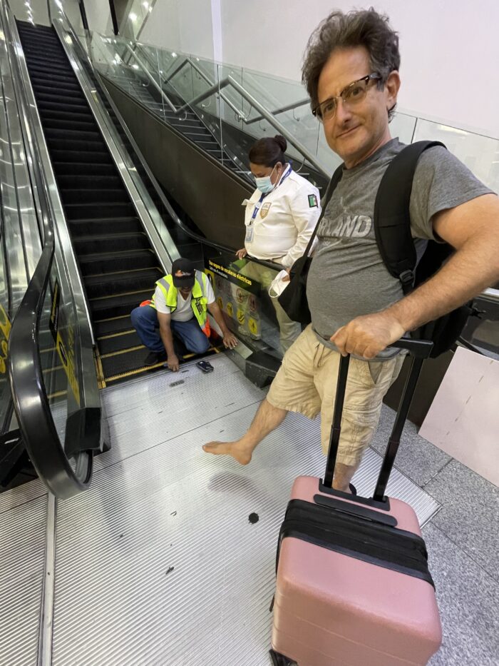 Traveler stands barefoot beside an escalator at Los Cabos International Airport as airport maintenance works to free his trapped slide.