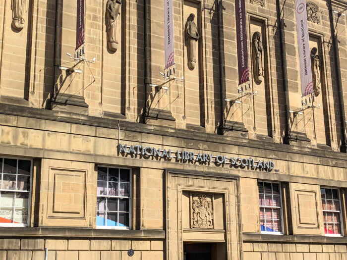 Exterior of the National Library of Scotland in Edinburgh, with historic stone architecture, sculpted figures and banners lining the façade.