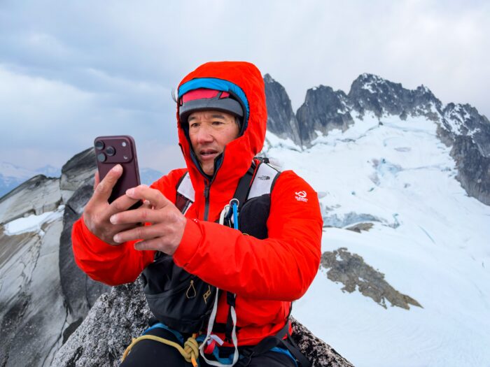 Phototgrapher Jimmy Chin in a bright red jacket uses an iPhone 18 while perched on a rocky alpine peak, with snow-covered mountains and glaciers in the background.