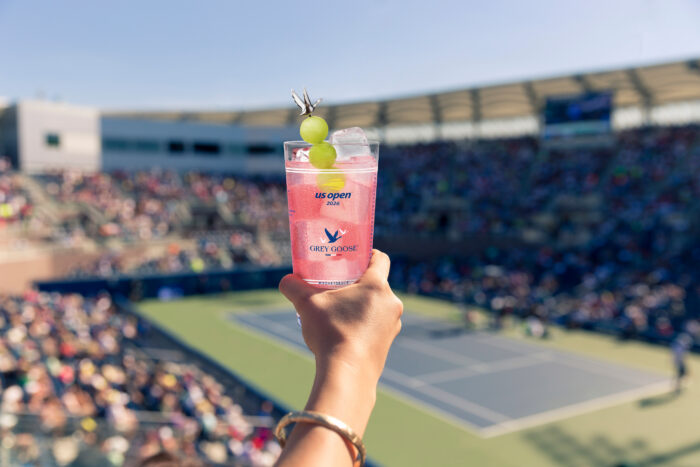 A hand holds a pink Grey Goose Honey Deuce cocktail garnished with honeydew melon balls overlooking a tennis court at the 2026 US Open.