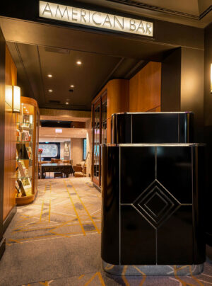 Entrance to the American Bar at The Savoy, featuring a black Art Deco host stand, warm wood paneling and geometric carpeting.