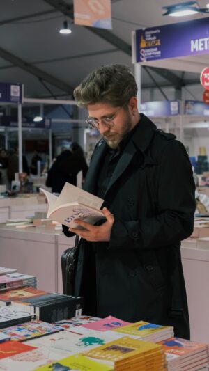Man in glasses browsing a book at a literary festival, surrounded by colorful books and publisher displays.
