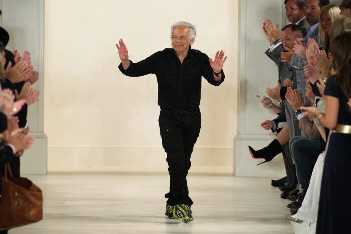 Fashion designer Ralph Lauren walks the runway in an all-black outfit and sneakers, waving to an applauding audience after a fashion show.