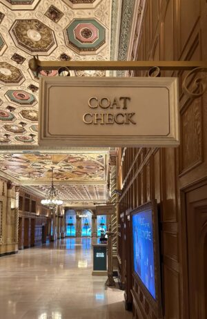 Ornate historic hallway with a hanging “Coat Check” sign beneath an intricately decorated ceiling.